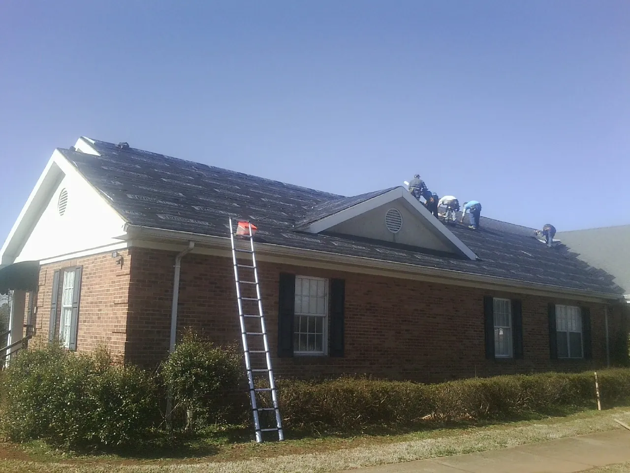 Crew tearing shingles off a brick ranch roof with a ladder set — Metal Roof Repair work by our crew in Tipp City