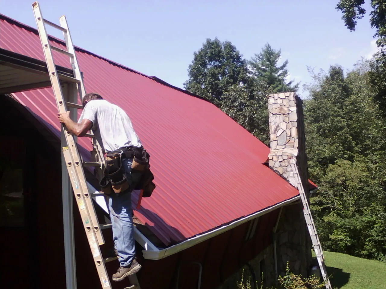 Roofer on a ladder beside a red metal roof and stone chimney — Soffit & Fascia Repair by Roofing Brothers in Tipp City